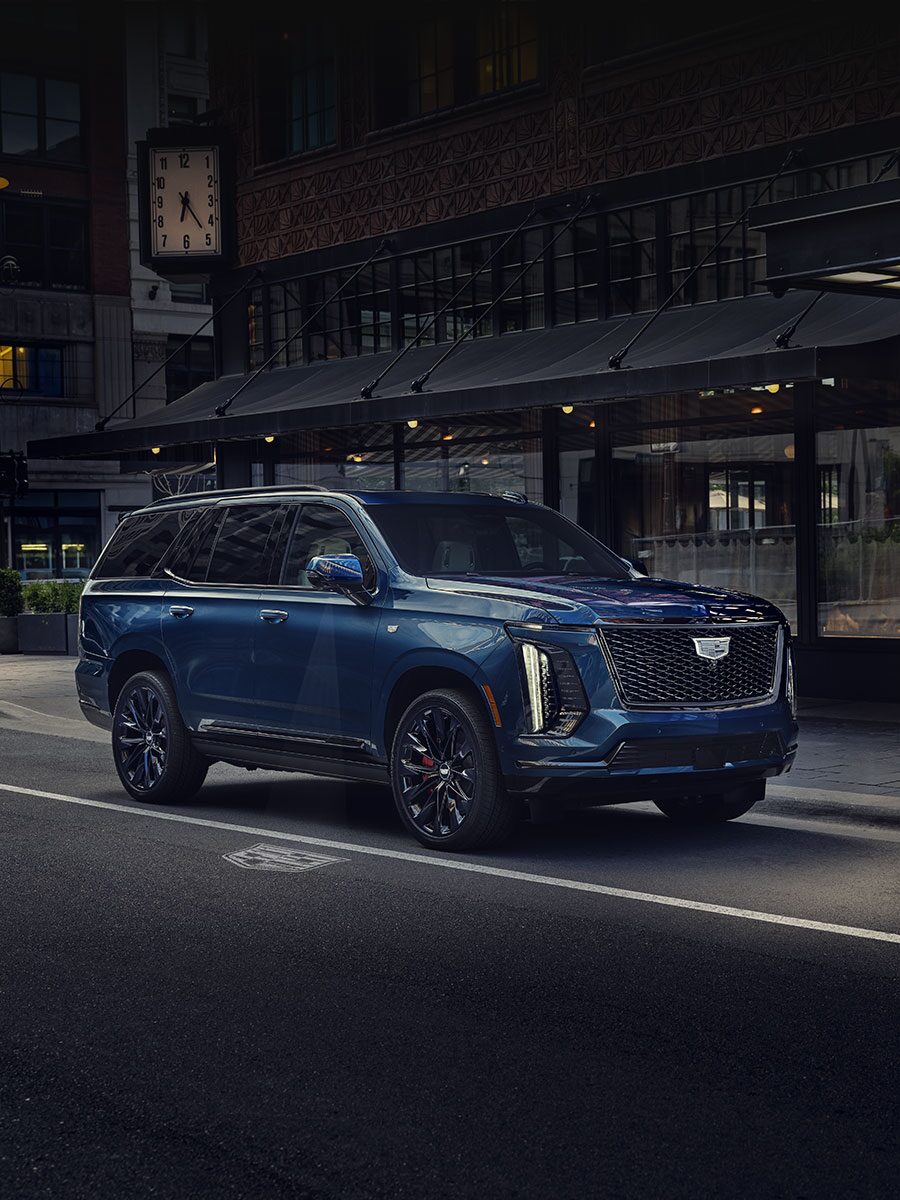 Un Cadillac Escalade 2027 bleu stationné dans une rue urbaine devant des immeubles du centre-ville, avec le texte « 2027 Escalade Features »