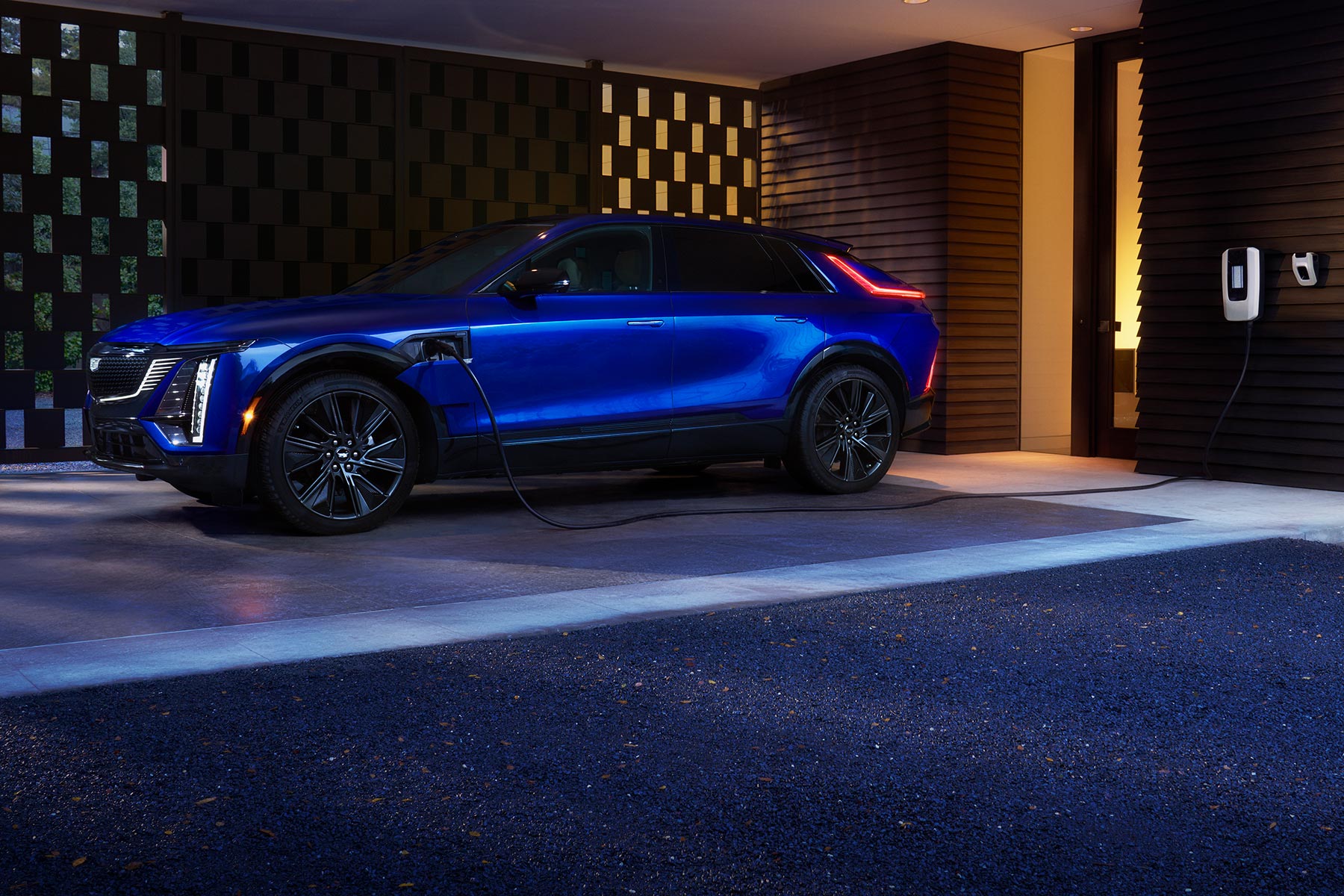 Side view of a blue 2027 Cadillac LYRIQ connected to a wall-mounted home charging station outside a modern residence at night.