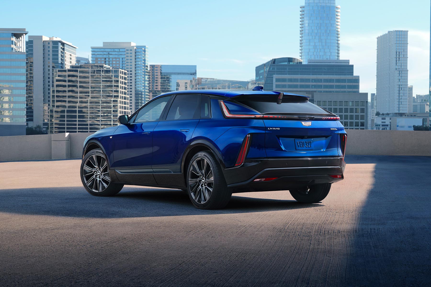 Rear three-quarter view of a blue 2027 Cadillac LYRIQ parked on a rooftop parking deck with a city skyline in the background.