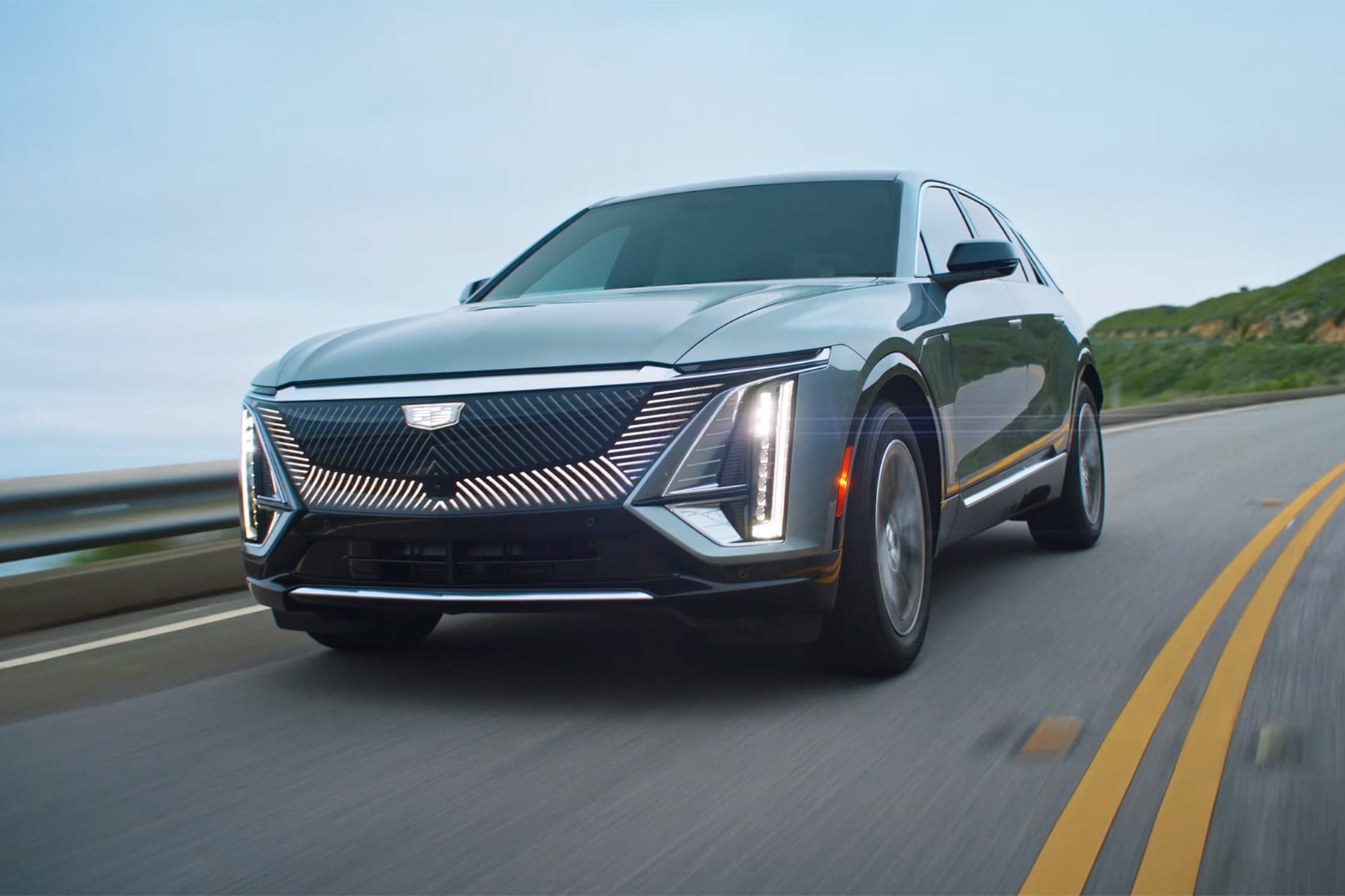 Front three-quarter view of a silver 2027 Cadillac LYRIQ driving along a coastal highway.