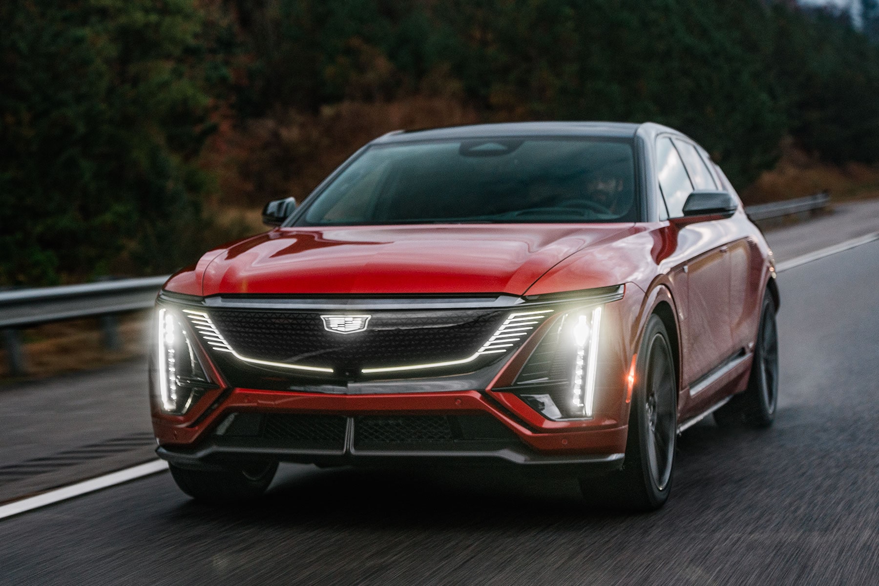 2027 Cadillac LYRIQ-V SUV driving alone on the road with its headlights on.