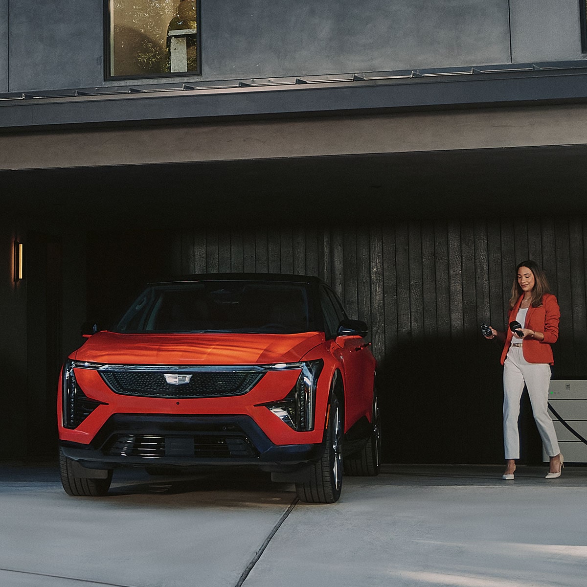 Une personne marchant vers une Cadillac OPTIQ rouge 2027 stationnée dans un garage avec un chargeur de VE à la main.