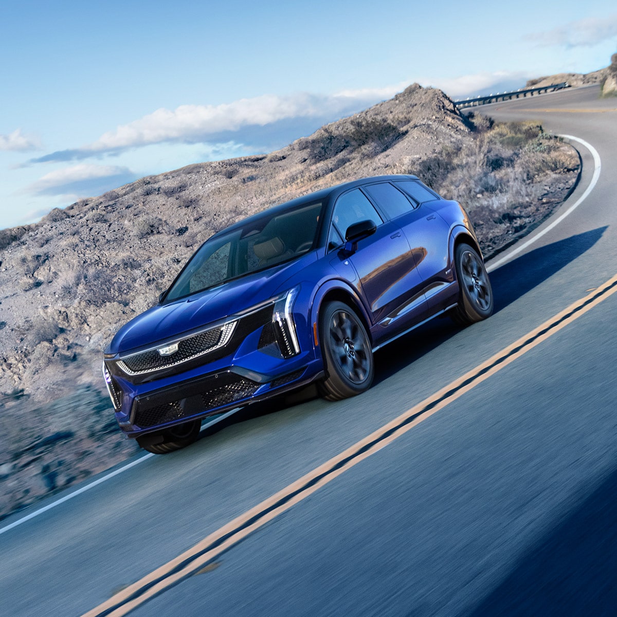Une Cadillac OPTIQ-V bleue 2027 roulant dans le virage d’une route de montagne.