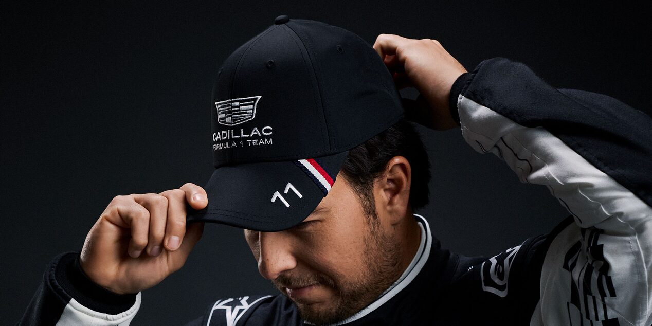 A Team Member Wearing A Black Cadillac-Branded Cap Adjusts the Brim While Posed Against A Dark Studio Background.