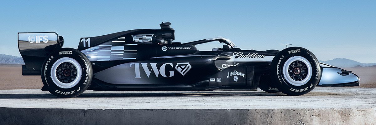 A Side Profile Of A Black-And-White Formula Race Car With Cadillac Branding Parked On A Sunlit Desert Flat With Mountains In the Background.