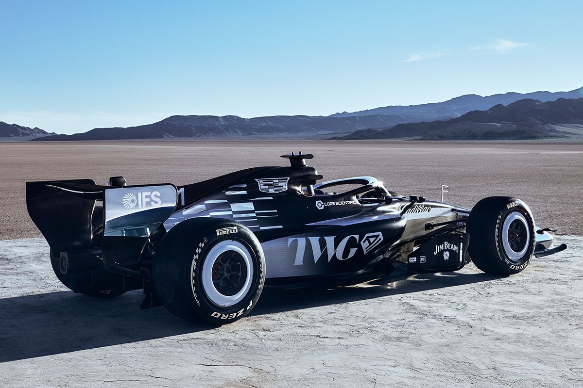 A Sleek Black-And-White Formula Race Car With Prominent Cadillac Branding Parked On A Sunlit Desert Flat With Mountains In the Distance.
