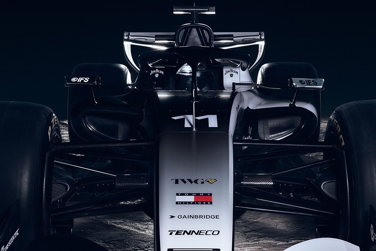 A Close Front View Of A Cadillac-Branded Formula Race Car Highlighting the Nosecone, Suspension Arms, And Aerodynamic Bodywork Against A Dark Backdrop.