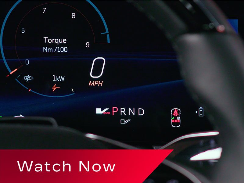 A Digital Cadillac OPTIQ Driver Display Highlighting Torque Readouts, Speed Information, And PRND Gear Indicators Partially Obscured By the Steering Wheel Above a Red Banner Reading “Watch Now.”