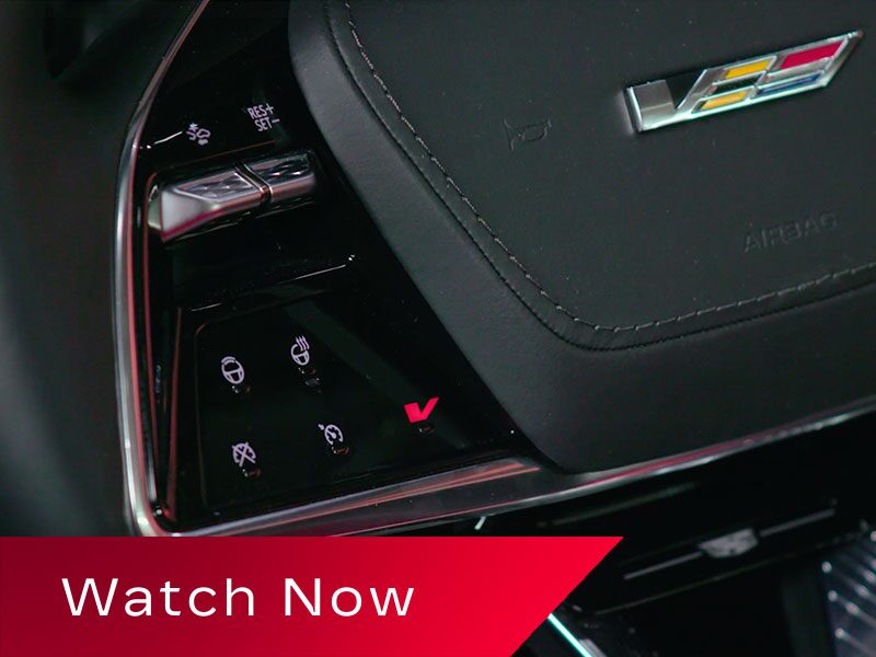 A Close-Up View Of A Cadillac OPTIQ Steering Wheel Featuring Illuminated Touch Controls And A Prominent V-Series Badge Above A Red Banner Reading “Watch Now.”