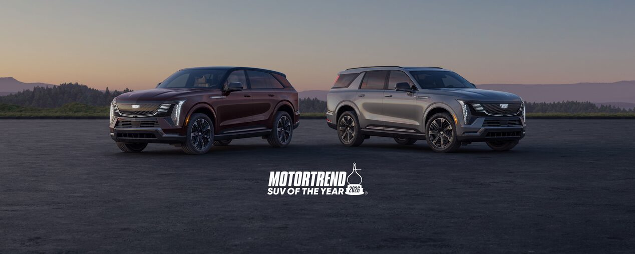 A Pair Of 2026 Cadillac Escalade IQ And IQL SUVs Are Parked On An Open Paved Surface At Sunset Above A Large MotorTrend SUV Of The Year 2026 Logo And Two Explore Model Buttons.