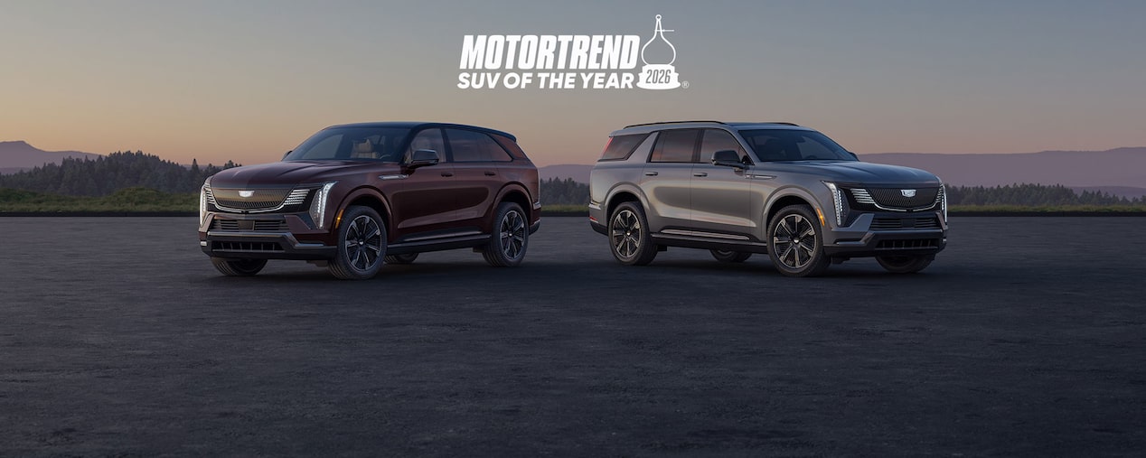 A Pair Of 2026 Cadillac Escalade IQ And IQL SUVs Are Parked On An Open Paved Surface At Sunset Above A Large MotorTrend SUV Of The Year 2026 Logo And Two Explore Model Buttons.