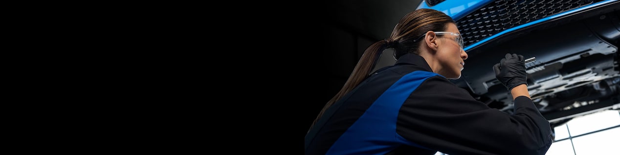 A Woman Certified Technician Looking Under a Lifted Vehicle in a Service Garage.