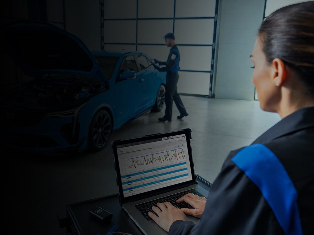 Cadillac Certified Service Technician Working on a Laptop with a Vehicle Nearby