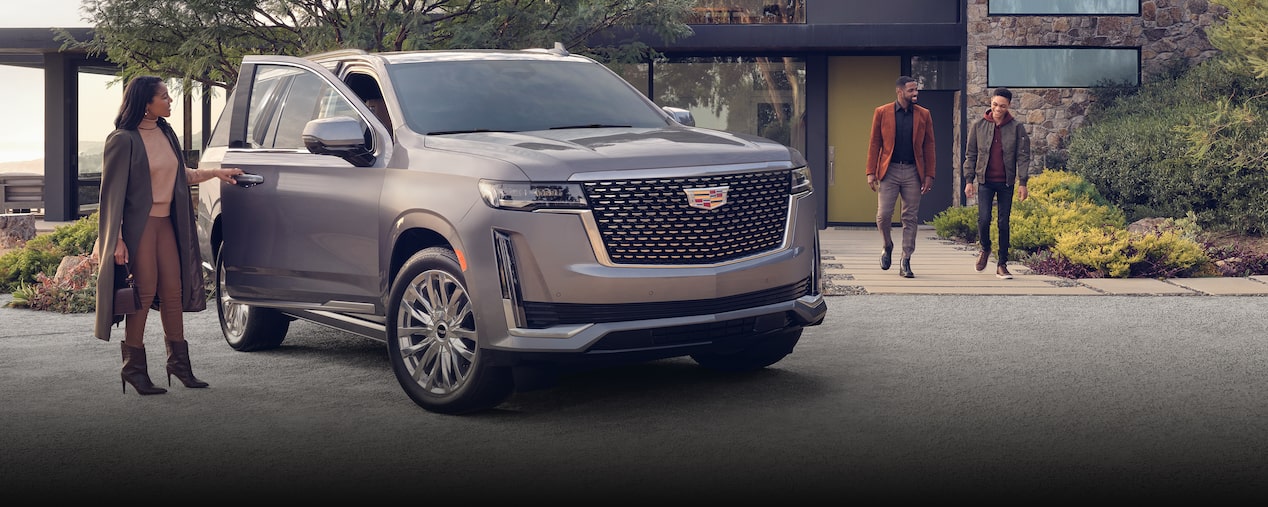 Cadillac Escalade sits in the driveway of a modern home. A woman stands at the open passenger side door while a man and a young boy walk out of the home toward the car. OnStar Insurance logo is in the bottom right corner.