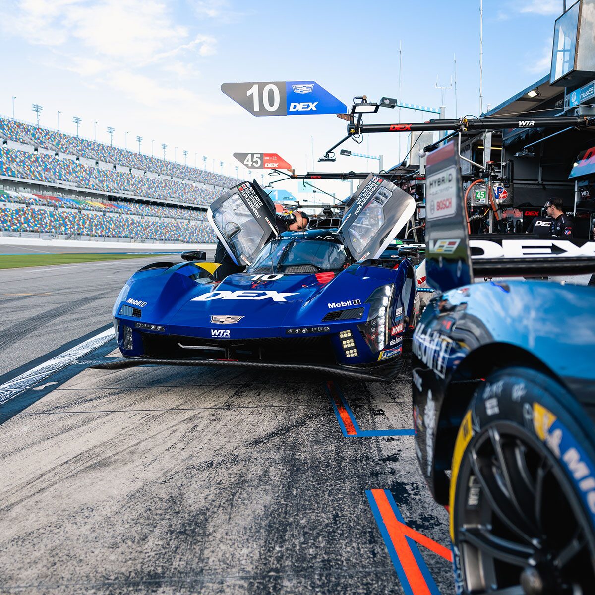 A Blue Cadillac V-Series.R Race Car Sits in the Pit Lane During a Stop as Crew Members Service the Vehicle Under Bright Daytime Light.