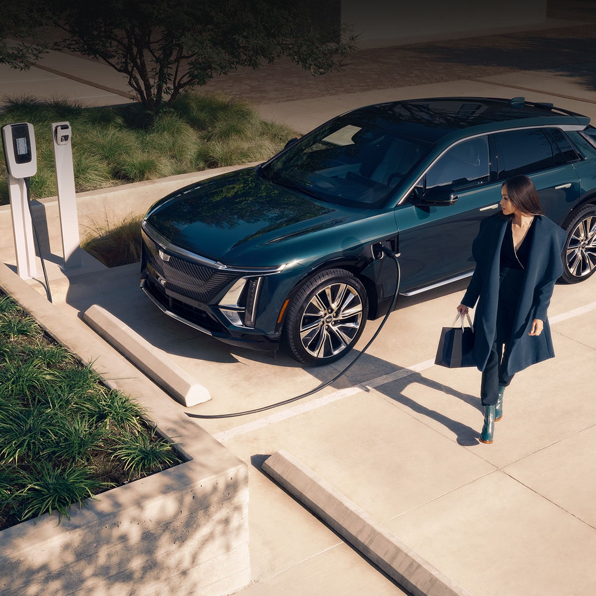 A Black Cadillac LYRIQ Is Parked At an EV Charging Station As a Woman Walks Away Along a Sunlit Sidewalk.