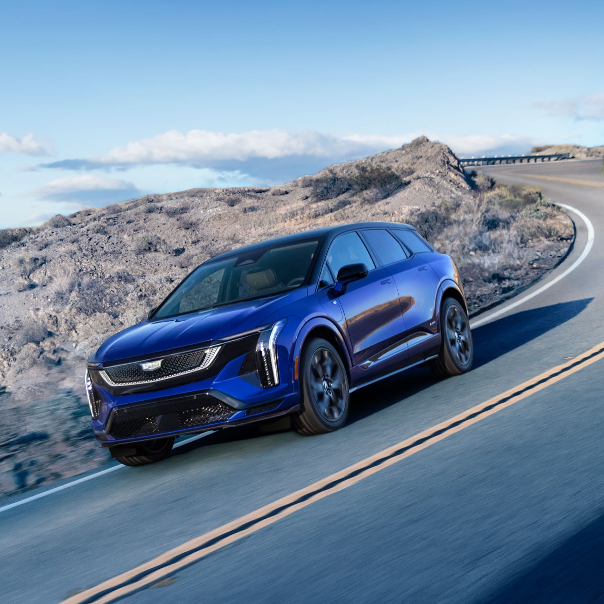Three-quarter View of a Blue Cadillac OPTIQ-V Driving Down the Curve of a the Road in a Rocky Desert